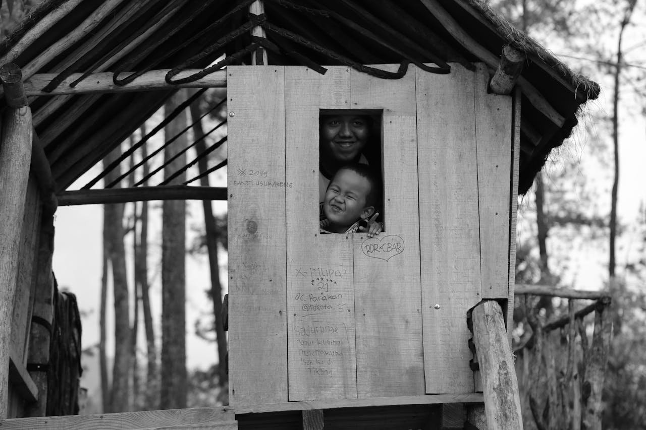 our-experience A playful moment captured as children peek from a wooden playhouse in a forest setting.