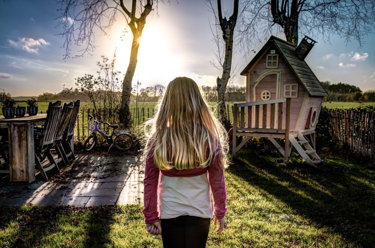 about-02 A child enjoying a sunny day in a backyard with a wooden playhouse and trees.