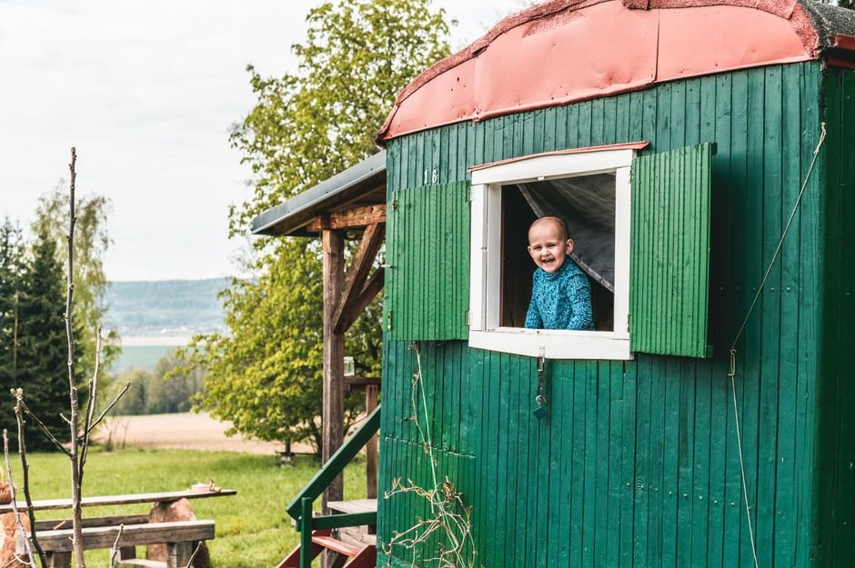Džiaugsmingas vaikas šypsosi pro žalio namelio langą gamtoje – puiki vieta žaidimams